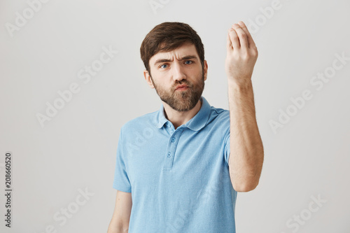 Cute bearded european male standing with raised hand and showing italian gesture and puckered lips, frowning, being serious while standing against gray background. Brother proves his point of view