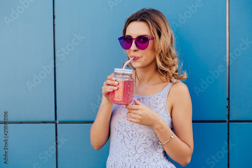 Foto Young trendy woman in sunglasses sipping fresh cold beverage, drinking through the straw, looking at camera, outdoors
