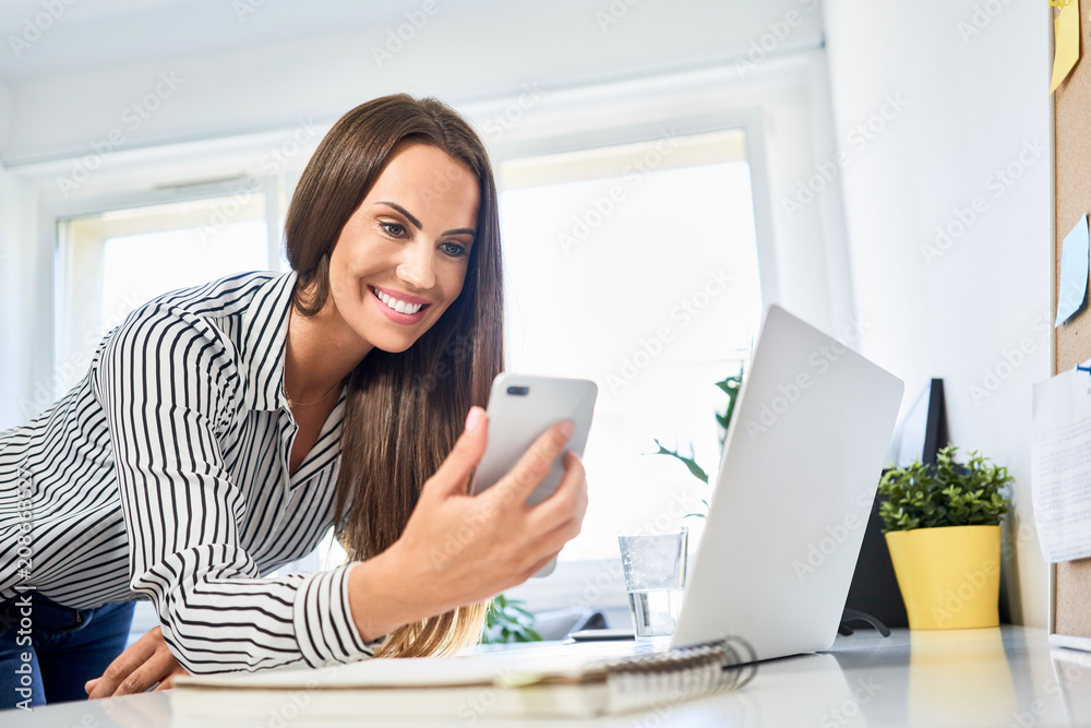 Woman looking at phone while leaning on desk with laptop in home office Woman looking at phone while leaning on desk with laptop in home office