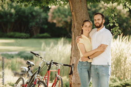 Young couple of cyclists is hugging outdoors. Young man and woman in love smiling and looking at camera in summer park. Loving couple on a date.