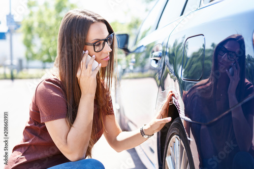 Yougn woman inspecting her scratched car and calling insurance agent