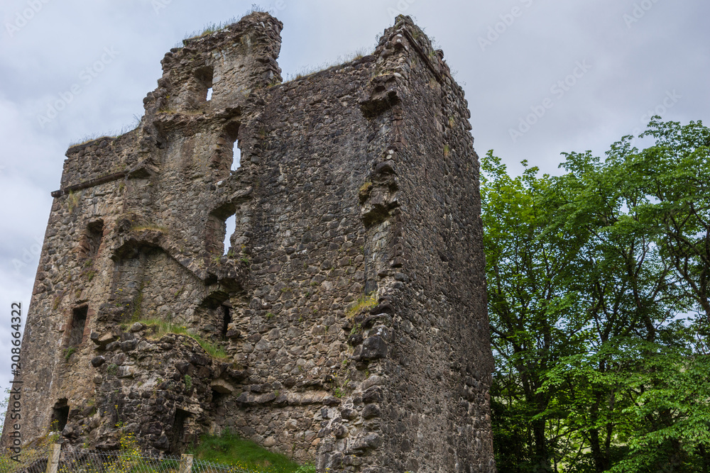 Invergarry, Scotland - June 11, 2012: Tall dark-brown ruined wall with window openings of castle Invergarry, set in green forest under light blue sky.