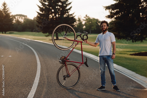 Handsome guy with bicycle resting outdoors. Young cheerful cyclists holding bicycle and looking at camera on country road.