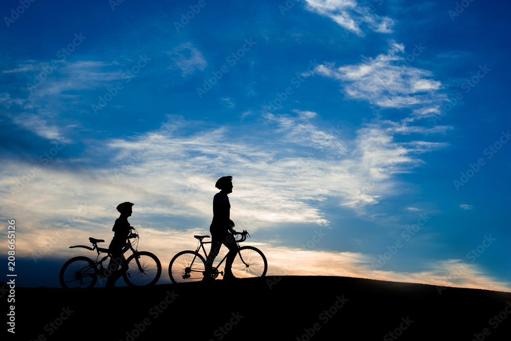 Young couple of cyclists walking at sunset. Young man and woman with bicycles at evening sky background. Amazing rest together outdoors. Young couple of cyclists walking at sunset. Young man and woman with bicycles at evening sky background. Amazing rest together outdoors.