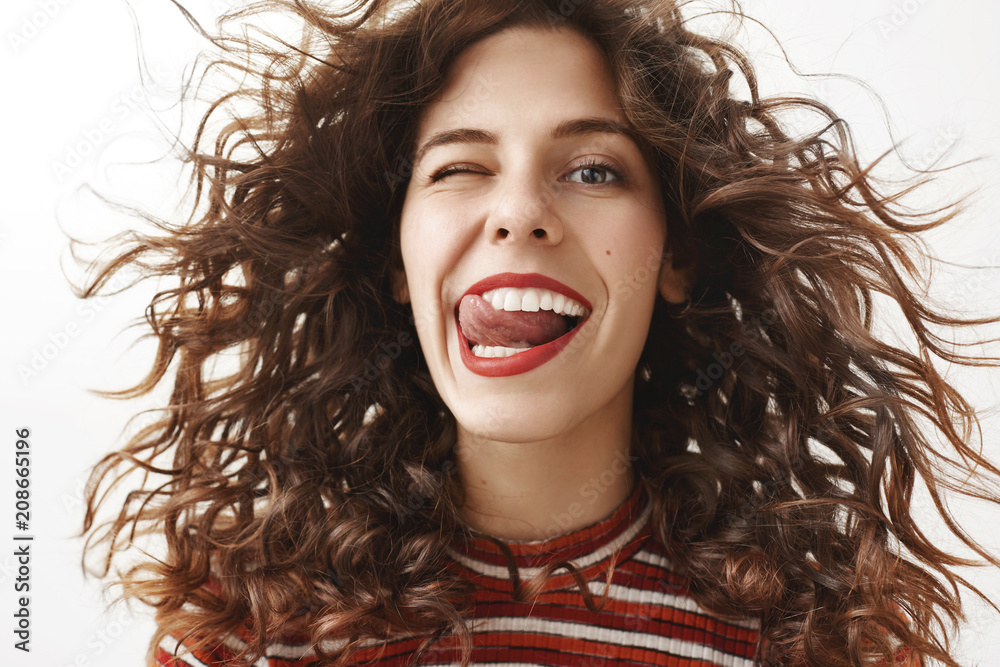 Close-up shot of carefree feminine and sensual european woman with curly hair that waves in air, sticking out tongue joyfully, smiling broadly while winking flirty at camera, being happy and confident Close-up shot of carefree feminine and sensual european woman with curly hair that waves in air, sticking out tongue joyfully, smiling broadly while winking flirty at camera, being happy and confident