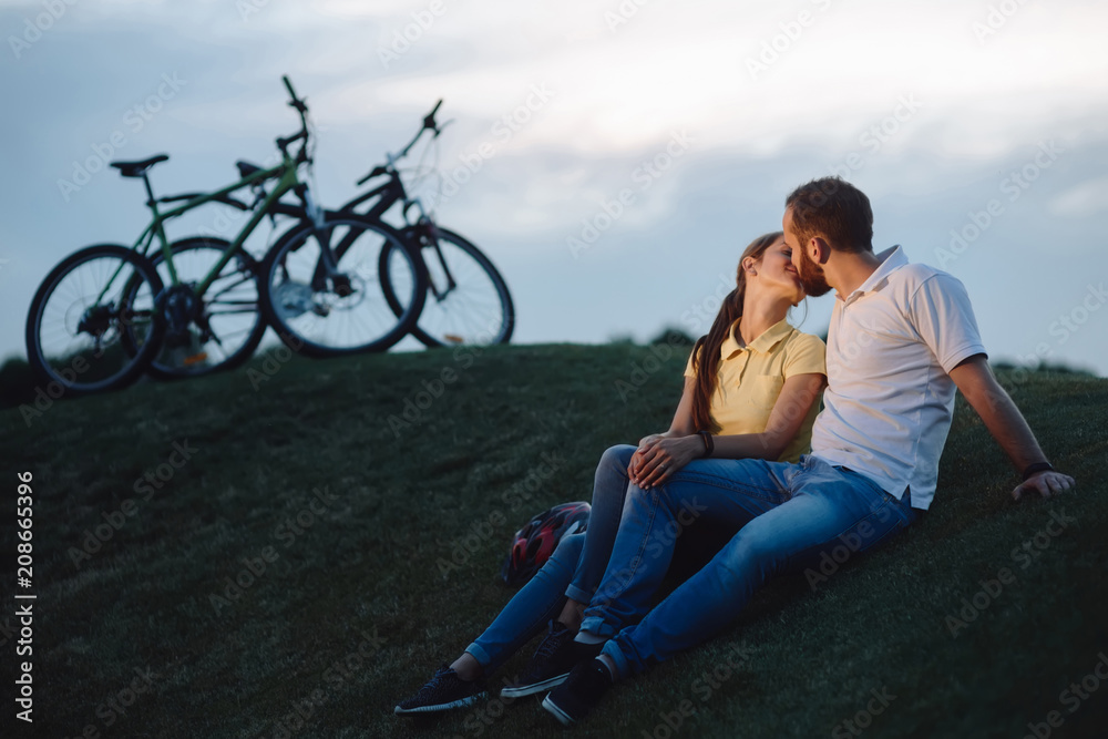 Romantic couple is kissing outdoors. Young man and woman in love is kissing during bike ride on green hill. Love and romance concept.