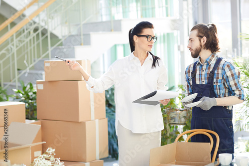 Serious beautiful female manager in white blouse holding clipboard with plan and explaining place for uploading to moving company employee unpacking box with plates