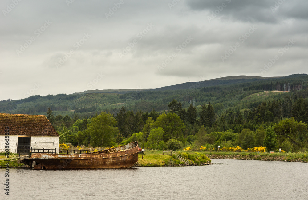 Fototapeta premium Fort Augustus, Scotland - June 11, 2012: Oich Canal behind the locks. Bucolic scenery with rusted boat wreck. Green hils with yellow flowers on horizon under heavy gray sky.