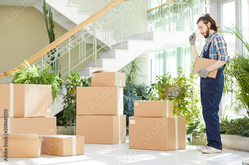 Serious pensive bearded young moving company employee with notepad concentrated on work counting cardboard boxes stacking in lobby