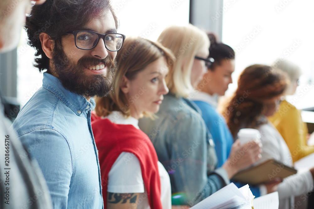 Cheerful excited handsome bearded man in denim shirt wearing eyeglasses standing in line and waiting for his turn while going to report to business forum Cheerful excited handsome bearded man in denim shirt wearing eyeglasses standing in line and waiting for his turn while going to report to business forum