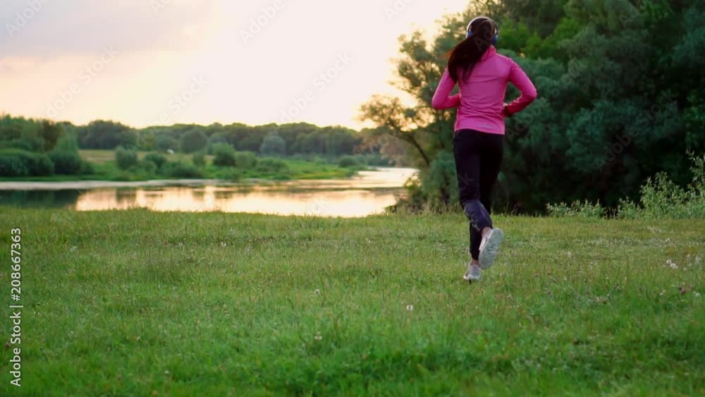 A girl in a pink jacket and black pants runs near the river in headphones preparing for the marathon A girl in a pink jacket and black pants runs near the river in headphones preparing for the marathon
