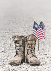 Old military combat boots with dog tags and two small American flags. Rocky gravel background with copy space. Memorial Day or Veterans day concept. Vintage tone.