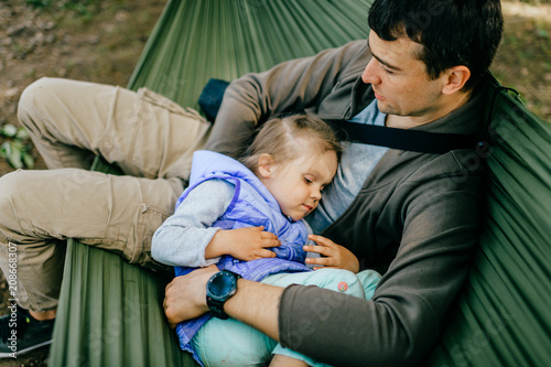 Modern Masculinity. Man in non-stereotypical role. Father caring his lovely daughter outdoor in summer.  Happy family expressing funny kind emotions at nature.  Parent with child leisure in hammock.