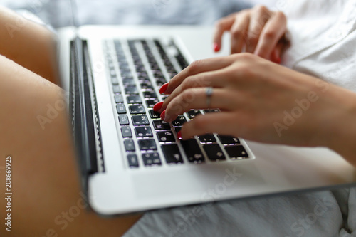 Hands close-up of a young girl in a man's shirt lying on the bed in the bedroom with a laptop on her lap and working. Work at home. The girl checks the mail in the morning on the tucked bed