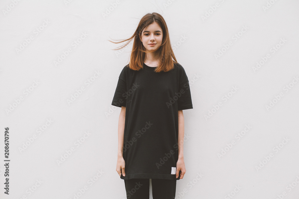 Young pretty girl wearing blank black t-shirt with space for your logo or design, mock-up of black cotton t-shirt, white wall in the background. Front view Young pretty girl wearing blank black t-shirt with space for your logo or design, mock-up of black cotton t-shirt, white wall in the background. Front view