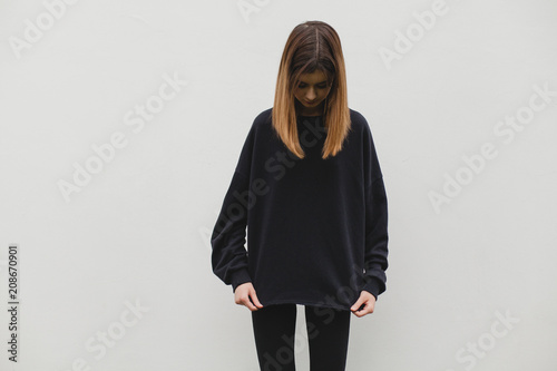 Young pretty girl wearing blank black hoodie with space for your logo or design, mock-up of black cotton sweatshirt, white wall in the background. Front view