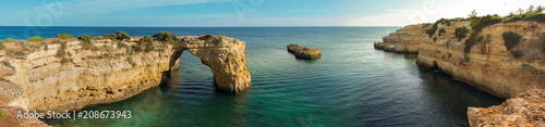 Panoramic view of the natural arch Arco  da Albandeira in the Algarve, Portugal. The arch and surrounding cliffs as well as a rock stump in the Atlantic ocean are shown on a sunny day with calm water. photo