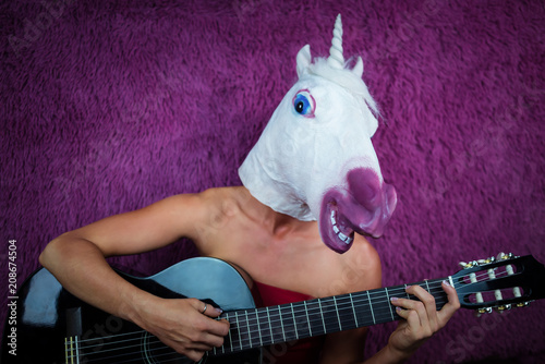 Freaky young woman in comical mask playing the guitar on the purple background. Portrait of unusual lady in red dress with musical instrument. Musical performance.