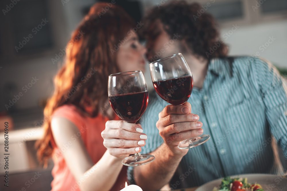 Focus on wineglasses in male and female hands. Affectionate young man and woman are kissing on background 
