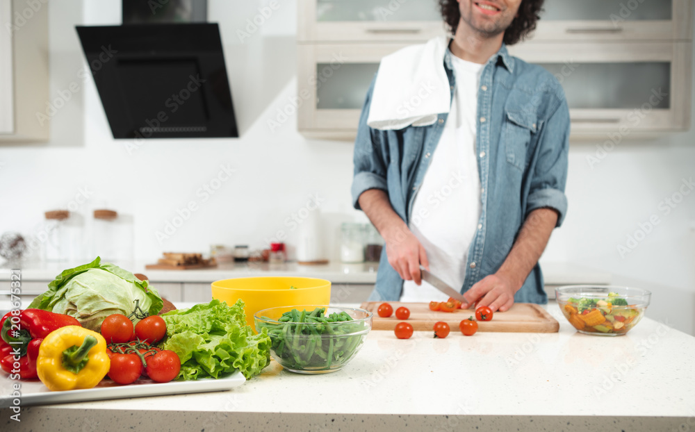 Cheerful young man is making cutting tomatoes while standing in kitchen. Focus on colorful vegetables om table 
