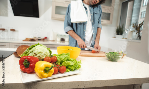 Close up focus on fresh peppers, tomato, lettuce and cabbage on the table. Young man is making salad on background 