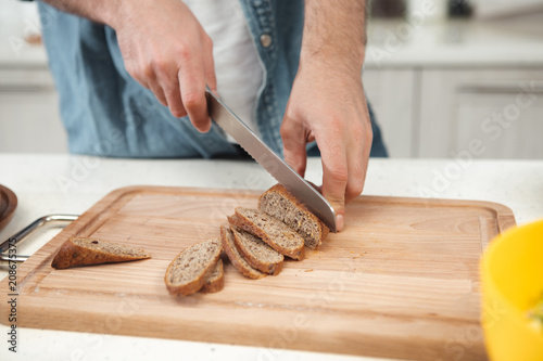 Close up of male hands cutting fresh bread with crust into thin pieces on wooden board. Man is holding knife while standing at table 