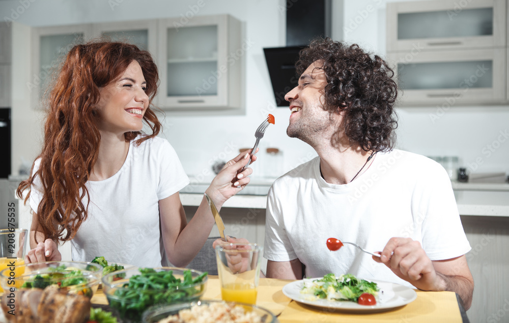 Taste this one. Joyful young woman is feeding her husband by a piece of tomato. She is laughing while sitting at table in cook room Taste this one. Joyful young woman is feeding her husband by a piece of tomato. She is laughing while sitting at table in cook room