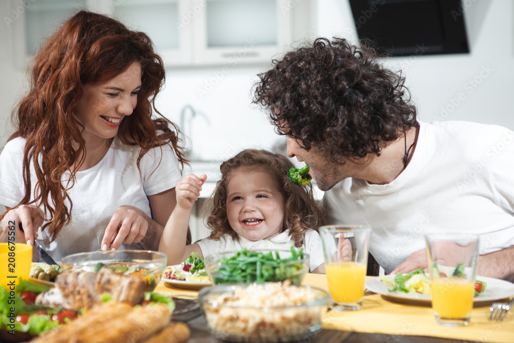 Happy childhood. Cheerful little girl is laughing while her father is keeping piece of broccoli near her. Mother is looking at them with love and smiling while eating salad Happy childhood. Cheerful little girl is laughing while her father is keeping piece of broccoli near her. Mother is looking at them with love and smiling while eating salad