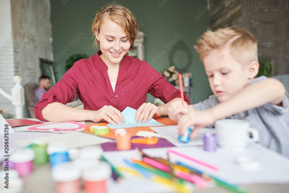 Adorable little boy and his pretty young mother wrapped up in making present for Fathers Day while gathered together at living room with stylish design Adorable little boy and his pretty young mother wrapped up in making present for Fathers Day while gathered together at living room with stylish design