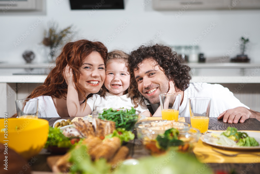 Friendly family. Portrait of happy little girl is hugging faces of her mom and dad. They are looking at camera and smiling while sitting at table in kitchen 