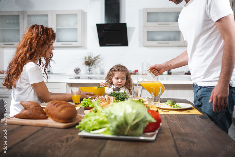 Caring mom and dad are feeding their child with healthy breakfast. Woman is holding a bowl with salad while man is pouring juice into glass. Pretty girl is looking at food with appetite Caring mom and dad are feeding their child with healthy breakfast. Woman is holding a bowl with salad while man is pouring juice into glass. Pretty girl is looking at food with appetite