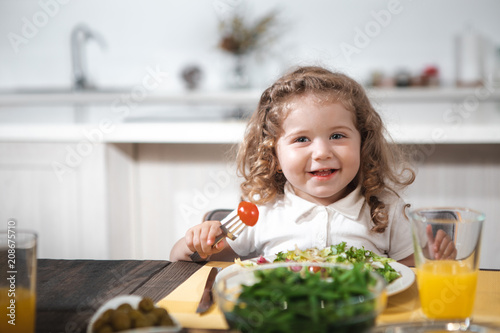 I like salad. Portrait of happy curly girl is eating vegetables with enjoyment. She is looking forward and smiling while sitting at table in kitchen 