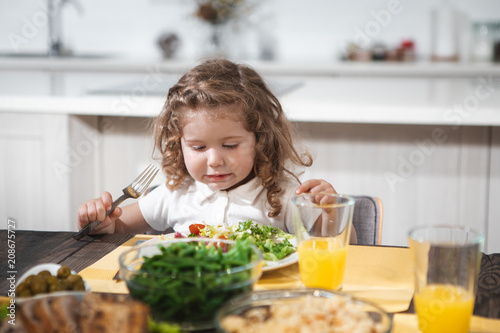 Portrait of pretty girl is looking into the plate at salad with interest. She is holding a fork while sitting at table in kitchen 