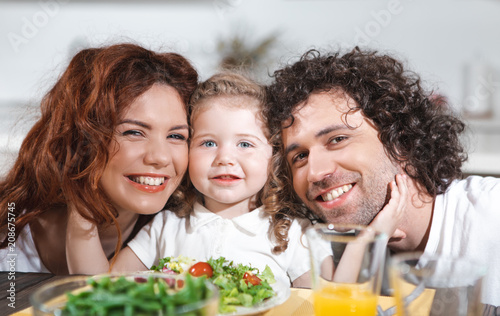Happy together. Portrait of glad father and mother are hugging their little daughter with love. They are eating salad in kitchen 