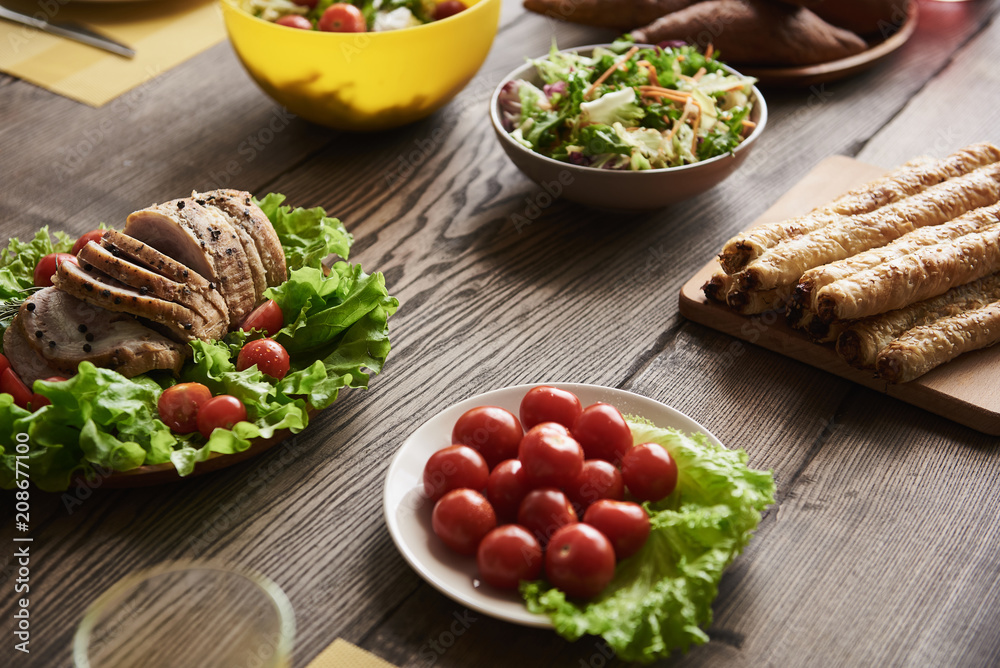 Close up of baked meat lying on lettuce together with white plate of cherry tomatoes. Bread and salads in bowls are nearby