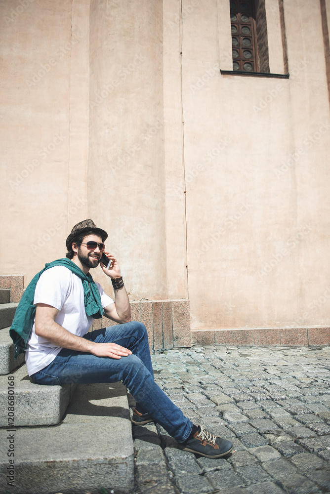 Connection concept. Happy man in trendy accessories taking seat on stairs outside. He is talking by mobile phone and smiling. Copy space in right side