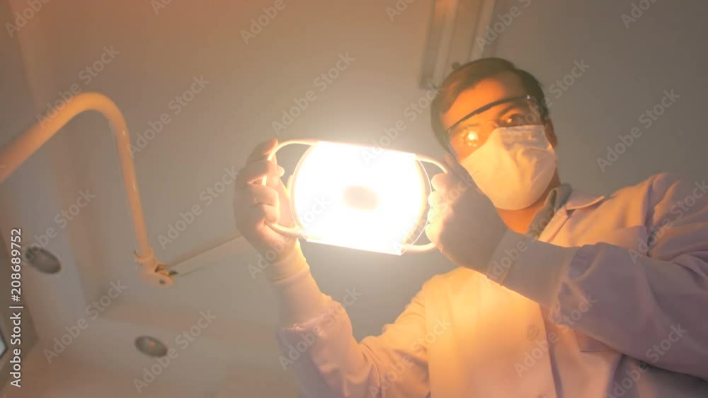 Caucasian dentist in mask turns the Light of dental lamp on, Lights into a patient's mouth, Patient point of view.