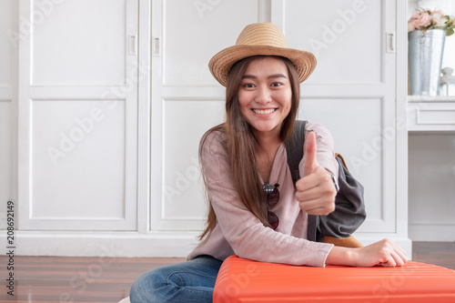 Young asian woman traveler thumbs up with orange suitcase prepare for holiday vacation at home.backpacker travel concept.