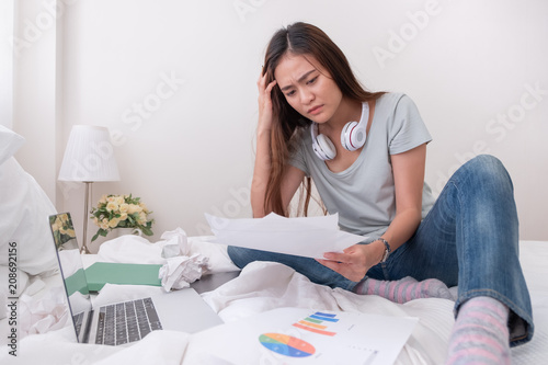 Asian woman freelancer headache about paperwork on bed with laptop in bedroom.work at home concept.stress working.