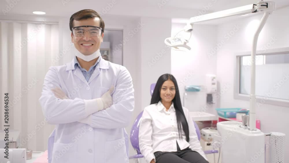 Portrait of smiling caucasian dentist in dental studio with young asian woman patient smiling in background. Portrait of smiling caucasian dentist in dental studio with young asian woman patient smiling in background.