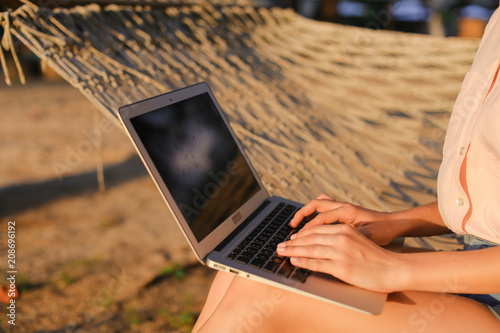 Close up female person using laptop and sitting on wicker hammock. Concept of modern technology and resting on beach. photo