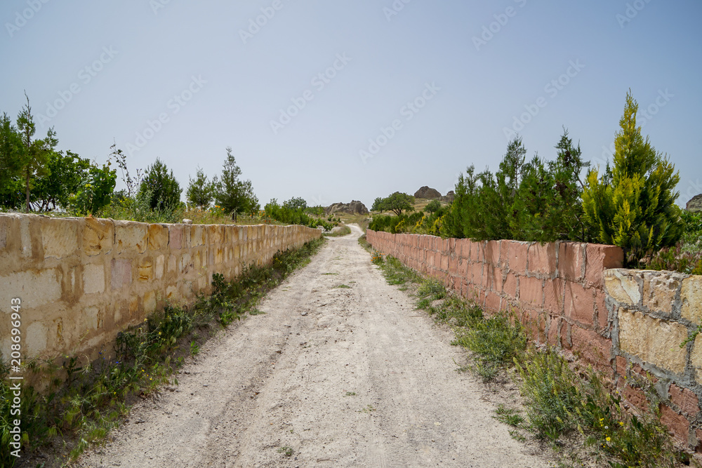 Walking unpaved rough sand road through stone brick low wall among landscape of dried and green ancient red valley with clear sky background, Cappadocia