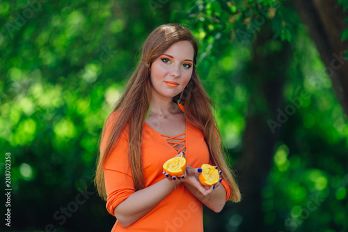 Portrait of pretty red hair woman with two halves of juicy delicious oranges at summer green park.