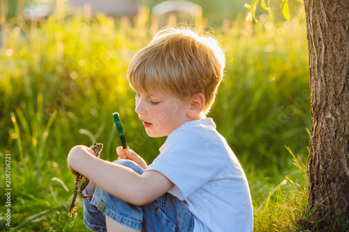 Cute preschooler blond boy looking at little frog in his hands through the magnifying glass at nature in sunset. Green background  side view. Childhood and researching concept.