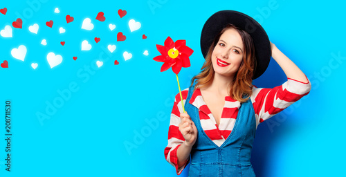 Portrait of young smiling red-haired white european woman in hat and red striped shirt with jeans dress with pinwheel on blue background with hearts