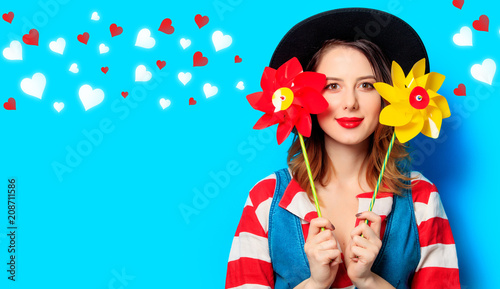 Portrait of young smiling red-haired white european woman in hat and red striped shirt with jeans dress with two pinwheels on blue background with hearts