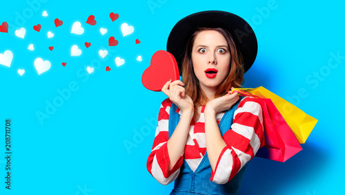 surprised red-haired white european woman in hat and red striped shirt with heart shape toy and shopping bags on blue background with hearts