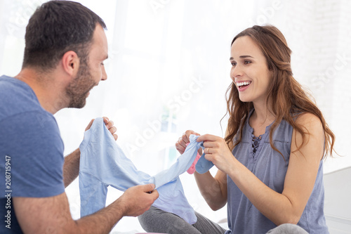 Clothes for baby. Cheerful pregnant woman and man carrying crawlers and talking