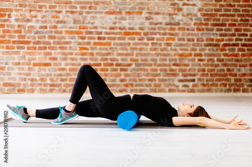 Young fitness woman in black sportswear doing stretching exercise with foam roller in modern loft interior at gym. Sport, fitness, lifestyle and people concept