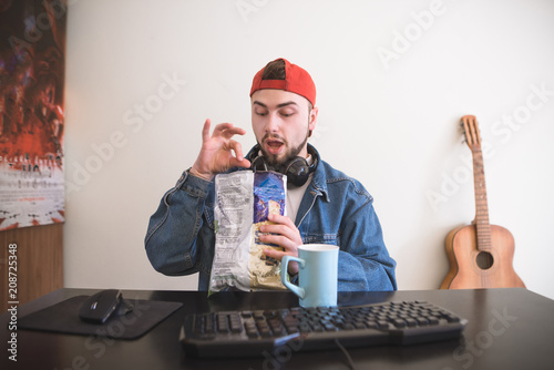 Portrait of a funny man with a beard and headphones who plays video games on a computer and eats a snack from a pack. Funny gamer eats chips while playing the computer